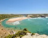 Atemberaubende Aussicht auf Praia de Odeceixe, wo ein Fluss auf den Ozean trifft. Grüne Hügel und ein weißes Dorf säumen die Küste.