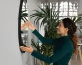 Mujer sonriente ajusta un espejo sin marco en la pared, mostrando cómo colgar un espejo sin marco para decorar su hogar.