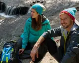 Para, co to jest trekking? Para w czapkach i kurtkach siedzi przy strumieniu, obok plecak.