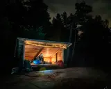 Person myser i ett upplyst vindskydd i skogen på natten. Ryggsäckar står utanför.