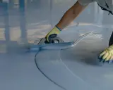 Persona con guantes amarillos aplica una capa de pintura azul clara sobre un suelo de terrazo con una llana.