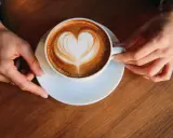 Eine Tasse Milchkaffee mit Herz-Latte-Art, gehalten von zwei Händen. Ein perfekter Start in den Tag, was ist Milchkaffee?