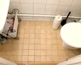 En liten toalett med beige kakelgolv. En tidningskorg står vid handfatet, och en toalettrulle och borste står bredvid toaletten. Kanske dags att måla kakel?
