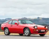 Roter Porsche 925 mit ausklappbaren Scheinwerfern steht auf einer Straße, im Hintergrund bewaldete Hügel.