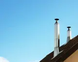 Dos tubos de chimenea metálicos se alzan sobre un tejado marrón contra un cielo azul claro. El sol calienta el metal, listo para aprovechar el calor.