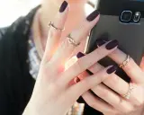 Dłonie z ciemnymi, **paznokciami półokrągłymi** i złotymi pierścionkami trzymają smartfon.