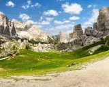 Un paesaggio mozzafiato nelle Dolomiti con le iconiche 5 torri che si ergono maestose. Un prato verde brillante invita al relax, con persone che riposano sotto il cielo azzurro.
