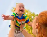 Niño con síndrome de Down, sonriente, es levantado por una persona. Sus rasgos son visibles y llenos de alegría.