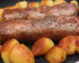 Deux andouillettes artisanales dorées, parsemées d'herbes, mijotent avec des pommes de terre rôties dans une poêle.