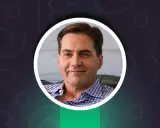 Uśmiechnięty Craig Wright w niebiesko-białej koszuli w kratę, z kluczem w lewym górnym rogu.