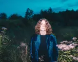 Młody mężczyzna z kręconymi włosami w aksamitnej, niebieskiej kurtce stoi pośród dzikich kwiatów i traw. Schafter ramotka w nocnej scenerii.