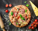 Gotowa do smażenia pizza fritta z salami, papryką i serem, obok pomidorki, ser i wałek do ciasta.