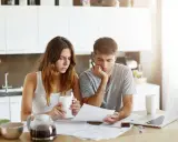 Couple pensif devant des papiers, peut-être une attestation d'assurance villégiature, dans une cuisine lumineuse.
