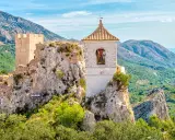 Das Castell de Guadalest thront auf einem Felsen, umgeben von grünen Bergen und dem blauen Meer in der Ferne. Ein Glockenturm ragt empor.
