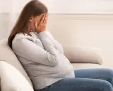 Kobieta w ciąży siedzi na kanapie, zakrywając twarz dłońmi. Jej postawa wyraża głęboki smutek, być może związany z diagnozą wady letalnej.