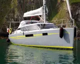 Le voilier "Bi-loup 109" gris avec des bandes jaunes, ancré près d'une falaise verdoyante.