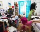 Chica en su habitación desordenada, hablando por teléfono, rodeada de ropa y objetos esparcidos.