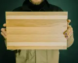 Hände halten ein rechteckiges Schneidebrett aus naturbelassenen holzbrettern mit hellen und dunkleren Streifen.