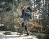 Mężczyzna w zimowej kurtce i czapce idzie przez las, wspierając się na kijach trekkingowych. Śnieg i suche liście pokrywają ścieżkę.