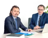 Dwóch mężczyzn w garniturach siedzi przy biurku z dokumentami i laptopem.