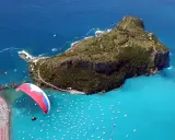 Parapendio su un mare turchese vicino a isole calabresi, con una spiaggia affollata e tante barche.