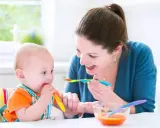 Uśmiechnięta mama karmi łyżeczką 7 miesięczne dziecko, które samo próbuje jeść.