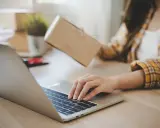 Osoba pakuje paczkę, sprawdzając na laptopie, kto ponosi koszty zwrotu towaru.