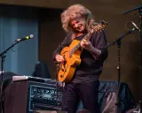Muzyk z burzą blond włosów gra na gitarze, tworząc wspaniały śpiew w duecie z niewidocznym wokalistą.