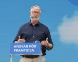 Man med glasögon och mikrofon talar vid ett podium med texten "Ansvar för framtiden". Hans gestik och budskap speglar en liberalism ideologi.