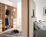 Un armario moderno y funcional, ideal para aprender como organizar un armario. Ropa colgada, cajones y estantes ordenados, con una bicicleta naranja y una cama al fondo.