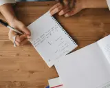 A person's hands are writing notes in a notebook. The notes mention "Theme: coronavirus", "self-isolation", and "lockdown".
