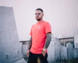 Kaz Belmondo w koralowej koszulce i z tatuażami na ramionach, stoi na tle betonowych bloków i falistej ściany.