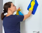Mujer con guantes amarillos y paño azul limpia puerta lacada en blanco con spray.