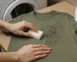 Manos frotando manchas negras en camiseta verde con esponja, cerca de una lavadora.