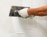 Mano enguantada aplicando estuco a una pared con una llana dentada. El proceso de estucar una pared requiere precisión.