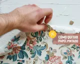 Mano retirando papel pintado floral de una pared blanca.