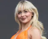 Blonde Frau mit voluminösen Blow-out-Haaren und einem strahlenden Lächeln in einem orangefarbenen Kleid.