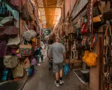 Bunte Taschen und Gürtel hängen in einem engen Gang des Marrakesch Basars. Ein Mann geht mit einer blauen Tüte durch das bunte Treiben.
