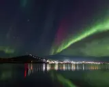 Gröna och lila norrsken dansar över en stjärnklar himmel ovanför Kiruna. Stadens ljus speglas i det lugna vattnet.