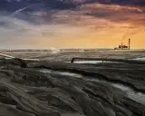 Krajobraz postindustrialny, gdzie rury odprowadzają odpady, a w oddali widać elektrownię. Woda i ziemia tworzą surowy obraz, symbolizujący proces rekultywacji.