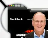 Uśmiechnięty mężczyzna w okularach, obok logo BlackRock. Larry Fink, jeden z założycieli BlackRock, na tle strony internetowej firmy.