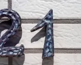 Dwa metalowe numery, 2 i 1, tworzą liczbę 21. Ich powierzchnia jest ciemnoniebieska z jaśniejszymi, nieregularnymi plamami, przypominającymi teksturę kamienia. Numery są zamontowane na białej, fakturowanej ścianie.