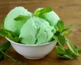Trois boules de glace verte menthe, rappelant la saveur de la glace Get 27, garnies de feuilles de menthe fraîche dans un bol blanc sur fond de bois.