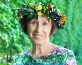 Eva Rydberg ler glatt med en blomsterkrans på huvudet, omgiven av grönska.