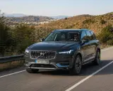 Volvo XC90 oscuro, uno de los suv mas fiables, circulando por una carretera rural con colinas verdes de fondo.