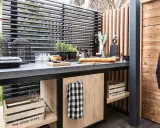 Cucina all'aperto fai da te con piano in cemento, pane, verdure e utensili. Un grembiule nero è appeso a una parete di legno.