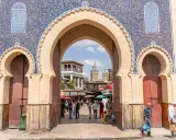 Prachtvolles Tor in Fes Stadt mit blau-goldenen Mustern. Menschen beleben den Platz dahinter.