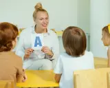 Nauczycielka pokazuje literę "A" grupie dzieci. Jedno z dzieci, 6-latek, uważnie słucha.