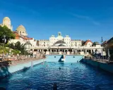 Soligt utomhuspool vid ett termalbad i Budapest. Människor solar och badar i det blå vattnet framför en imponerande byggnad.