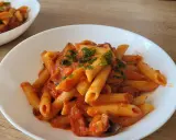 Penne z sosem pomidorowym i kawałkami kiełbasy, posypane natką pietruszki. Pyszny makaron z kiełbasą na białym talerzu.